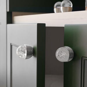 K&ouml;nnte beinhalten: Nahaufnahme von Schrankt&uuml;ren mit dekorativen Glas- und Metallkn&ouml;pfen. Die Kn&ouml;pfe haben eine klare, blasenartige Mitte und eine gerippte, silberfarbene Metallbasis. Die Schrankt&uuml;ren sind dunkelgrau und gr&uuml;n.