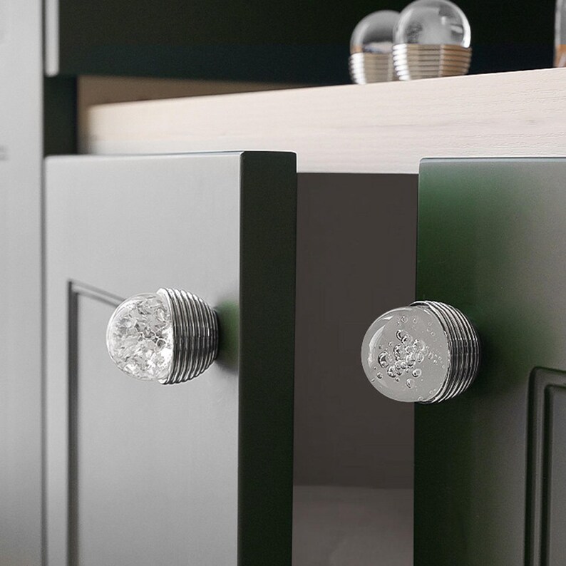 K&ouml;nnte beinhalten: Nahaufnahme von Schrankt&uuml;ren mit dekorativen Glas- und Metallkn&ouml;pfen. Die Kn&ouml;pfe haben eine klare, blasenartige Mitte und eine gerippte, silberfarbene Metallbasis. Die Schrankt&uuml;ren sind dunkelgrau und gr&uuml;n.