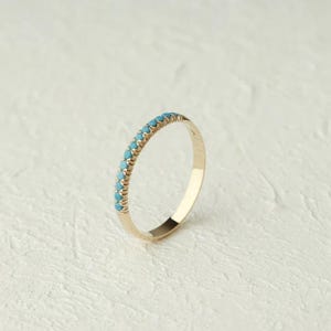 May include: A gold ring featuring a row of small, turquoise gemstones. The ring is set against a textured, off-white backdrop, highlighting the ring's design and the contrast of colours.