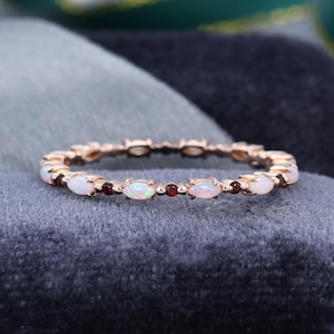 May include: Rose gold ring with alternating oval opals and small red gemstones. The ring is displayed on a dark grey surface. The design highlights the contrast between the white opals and the red gemstones.