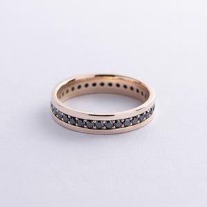 May include: A gold ring featuring a continuous row of small, black gemstones. The ring is displayed on a white surface, highlighting the contrast between the gold and black. The ring's band is approximately 0.64 cm wide.