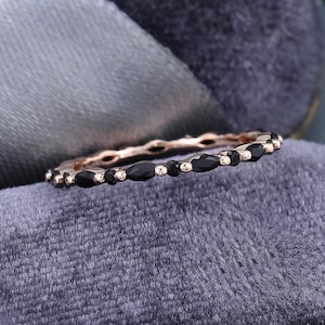 Peut inclure: Une bague en or rose ornée d'une rangée alternant des pierres noires ovales et rondes. La bague est présentée sur un fond de velours gris, soulignant le contraste des couleurs et des textures. Le design de la bague est délicat et élégant.