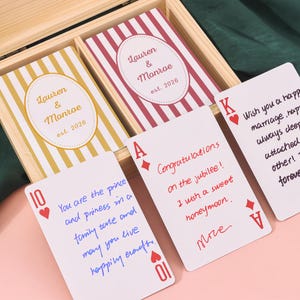 May include: A wooden box holds personalized playing cards. The cards feature the names "Lauren & Monroe" and the year "est. 2026". Some cards have handwritten messages, including "Congratulations on the jubilee!" and "I wish a sweet honeymoon."