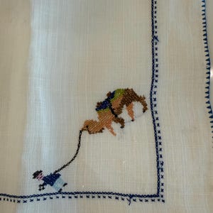May include: White linen handkerchief with a cross-stitched design featuring a person leading a camel. The camel is brown and tan with a green and blue saddle. The border is detailed with blue cross-stitch.