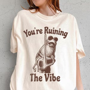 Puede incluir: Camiseta color crema con el texto "You're Ruining The Vibe" en marrón. La camiseta presenta una ilustración marrón de una rana con gafas de sol haciendo una peineta. La camiseta tiene mangas cortas y cuello redondo.