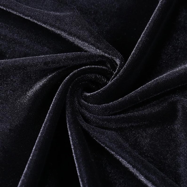 May include: A deep black velvet fabric featuring a dense, soft pile texture. The material is gathered in the center, creating natural folds and highlights that emphasize the light-reflecting quality of the dark, plush surface of the textile.