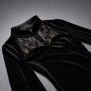 May include: A black long-sleeved top made of velvet fabric featuring a high collar and a decorative panel of black floral lace across the chest. The lace section is accented with a vertical row of four small, round, black buttons down the center.