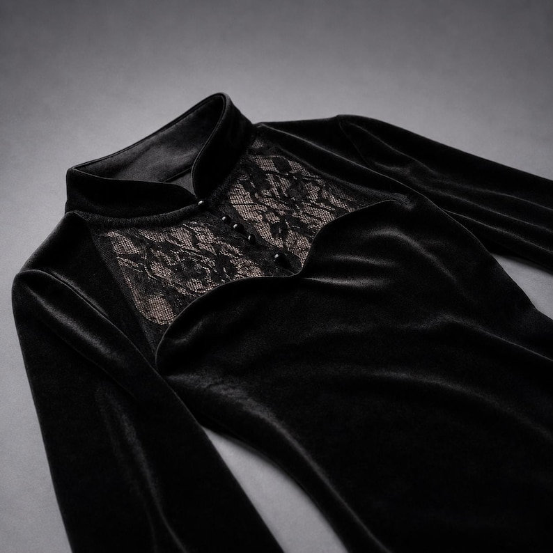 May include: A black long-sleeved top made of velvet fabric featuring a high collar and a decorative panel of black floral lace across the chest. The lace section is accented with a vertical row of four small, round, black buttons down the center.