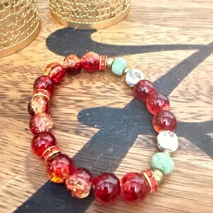 May include: A beaded bracelet featuring large, translucent red beads interspersed with smaller gold and patterned white beads. The bracelet also includes light green and gold accents, creating a colorful and eye-catching accessory.