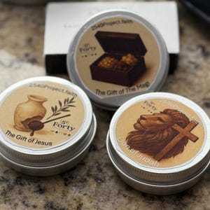 Könnte beinhalten: Drei kleine, runde Metalldosen mit beigen Etiketten. Die Etiketten zeigen Illustrationen und den Text: "The Gift of The Magi", "The Gift of Jesus" und "Lion of Judah". Jede Dose trägt den Text "2540Project.faith" und "Forty."