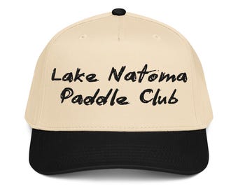 May include: A beige baseball cap with a black brim and a black button on top. The front of the cap features the black text Lake Natoma Paddle Club in a brushstroke font style. The cap has a structured crown and a curved visor.