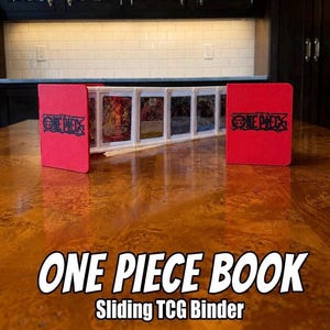 May include: Red "ONE PIECE BOOK" sliding TCG binder with the "ONE PIECE" logo. The binder holds several trading cards in protective cases. The binder is on a wooden table with a white brick wall in the background.