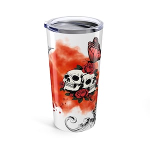 Puede incluir: Un vaso de viaje blanco con un diseño de dos calaveras, rosas rojas y una mariposa. El diseño incluye un fondo rojo en forma de nube y adornos decorativos. El vaso tiene una tapa plateada y tiene una capacidad de aproximadamente 590 ml.