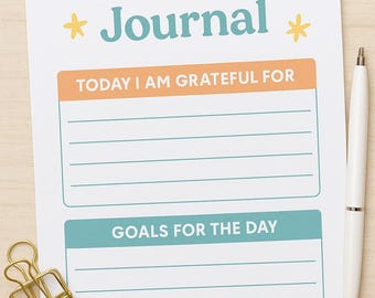 Może przedstawiać: Biały dziennik wdzięczności z napisem "Gratitude Journal" w kolorze turkusowym i pomarańczowym. Dziennik zawiera sekcje na codzienną wdzięczność i cele, ze złotą spinaczem i białym długopisem.