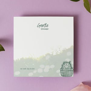 Peut inclure: Un bloc-notes carré avec le mot "Gentle" et une illustration de cactus sur un fond clair, sur un fond violet. Une fleur violet clair, des feuilles vertes et une bobine de ruban rose complètent la composition.