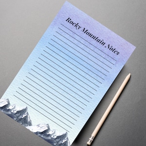 May include: A notepad with a watercolour design of a mountain range and the text "Rocky Mountain Notes". A wooden pencil lies beside the notepad. The notepad has a gradient of blue and purple. The notepad has lined pages.