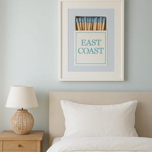 May include: A framed print featuring the words "EAST COAST" in light blue, above a depiction of matches. The artwork hangs above a bed with a white pillow and duvet. A wicker lamp sits on a wooden bedside table.