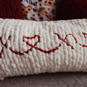 May include: A white, rectangular pillow with a textured surface. The word "XOXO" is embroidered in red across the front, with the "O"s replaced by hearts. The pillow is a decorative item.