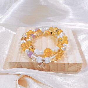 Puede incluir: Dos pulseras de cuentas con piedras preciosas amarillas, blancas y moradas. Una pulsera presenta un dije dorado con forma de cola de pez. Las pulseras se exhiben sobre un pequeño bloque de madera.