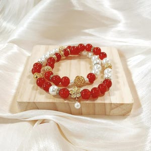 May include: Two beaded bracelets. One bracelet features red beads and gold accents, while the other has white pearls and gold details. A gold flower charm with a dangling pearl and a gold rose charm are also visible.