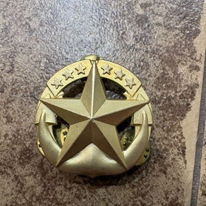 Puede incluir: Insignia militar dorada con una gran estrella de cinco puntas en el centro, rodeada por un círculo de estrellas más pequeñas y dos emblemas de ancla. La insignia tiene un acabado metálico y probablemente está hecha de latón o aleación similar.