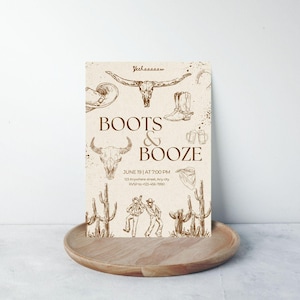 May include: A rustic invitation with the text "Boots & Booze" and cowboy-themed illustrations. The design includes images of a longhorn skull, cowboy boots, and cacti. The invitation is on a wooden tray.