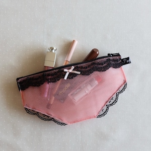 May include: A pink mesh zippered pouch shaped like underwear, featuring black lace trim along the top and bottom edges, a small light pink bow at the center, and a black zipper pull. The pouch contains three cosmetic items including a lipstick.