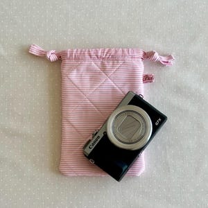 Può includere: Una custodia per fotocamera a righe rosa e bianche con chiusura a coulisse e una fotocamera Canon G7X nera. La borsa è trapuntata con un motivo a rombi. La borsa ha una piccola etichetta rosa con "Puf".