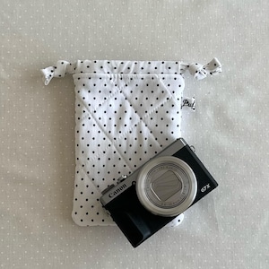 May include: A black Canon G7X camera rests on a white, quilted camera pouch with black polka dots. The pouch is closed with a drawstring. The background is a white fabric with small white polka dots.