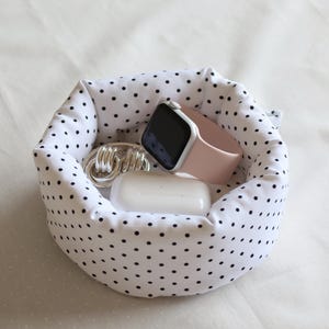 Puede incluir: Una cesta de tela blanca con lunares negros, que contiene un llavero plateado, una funda blanca para auriculares inalámbricos y un reloj inteligente rosa. La cesta es redonda y está sobre una superficie blanca.
