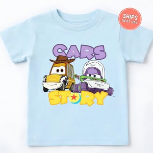A light blue t-shirt with a graphic design featuring two cartoon cars, one yellow and one purple, along with the text "CARS STORY".