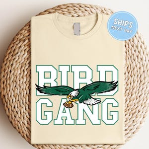 A beige t-shirt with a graphic design featuring a green eagle holding a football, along with the text "BIRD GANG" in large letters.