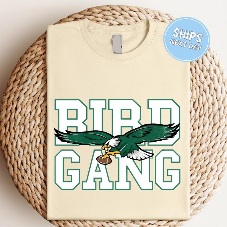 A beige t-shirt with a graphic design featuring a green eagle holding a football, along with the text "BIRD GANG" in large letters.