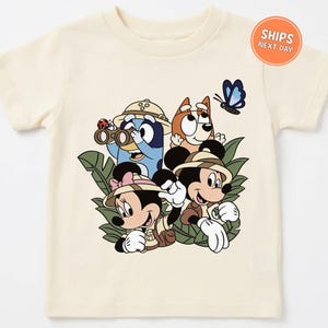 May include: A cream-colored short-sleeve t-shirt featuring a graphic print of Bluey, Bingo, Mickey Mouse, and Minnie Mouse dressed in safari gear. The characters are surrounded by green leaves, a ladybug, and a blue butterfly in the jungle.