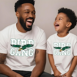 A man and a young boy wearing white t-shirts with the word "BIRD GANG" printed on them, both smiling and laughing.