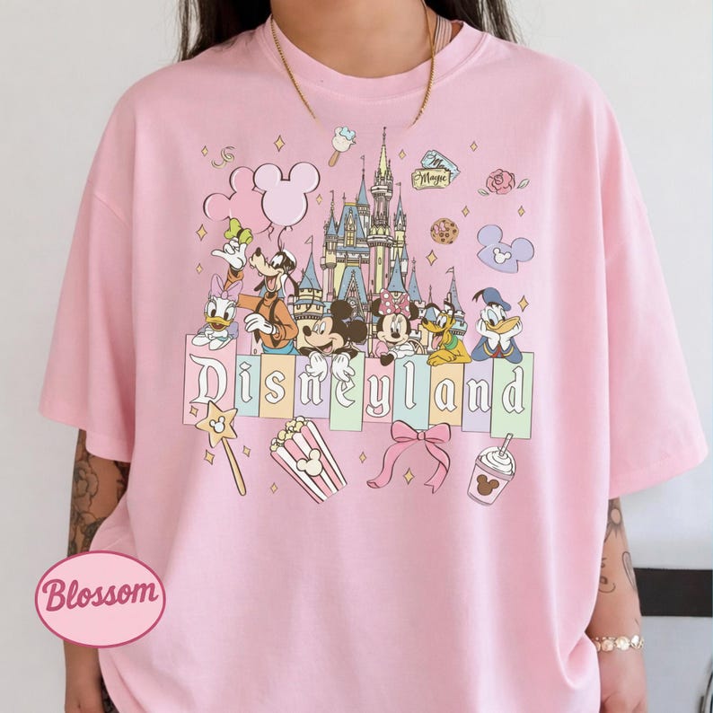 A pink t-shirt with a Disney theme graphic, featuring various Disney characters and landmarks such as the castle, Mickey Mouse, Minnie Mouse, Donald Duck, and other iconic Disney symbols.