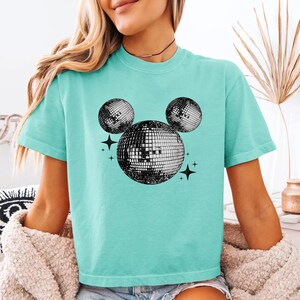 A woman wearing a turquoise t-shirt with a graphic of a disco ball and stars on it.