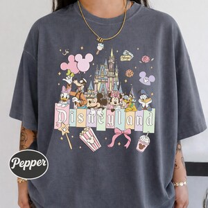 A person wearing a gray t-shirt with a Disney-themed graphic design featuring Mickey Mouse, Minnie Mouse, Donald Duck, and other Disney characters, along with the word "Disneyland" prominently displayed.