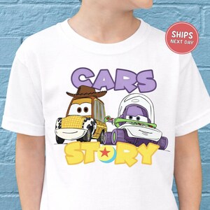 A white t-shirt with a graphic design featuring two cartoon cars, one with a cowboy hat and the other with a spacesuit, along with the text "CARS STORY".