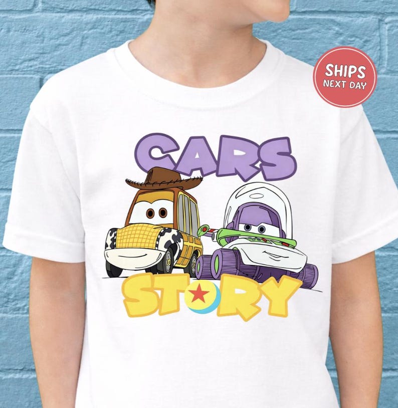 A white t-shirt with a graphic design featuring two cartoon cars, one with a cowboy hat and the other with a spacesuit, along with the text "CARS STORY".