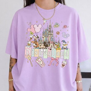 A person wearing a purple t-shirt with a Disney theme graphic, featuring various Disney characters and landmarks such as the castle, Mickey Mouse, Minnie Mouse, Donald Duck, and other iconic Disney symbols.