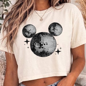 A woman wearing a white crop top with a graphic of two disco balls and stars on it.