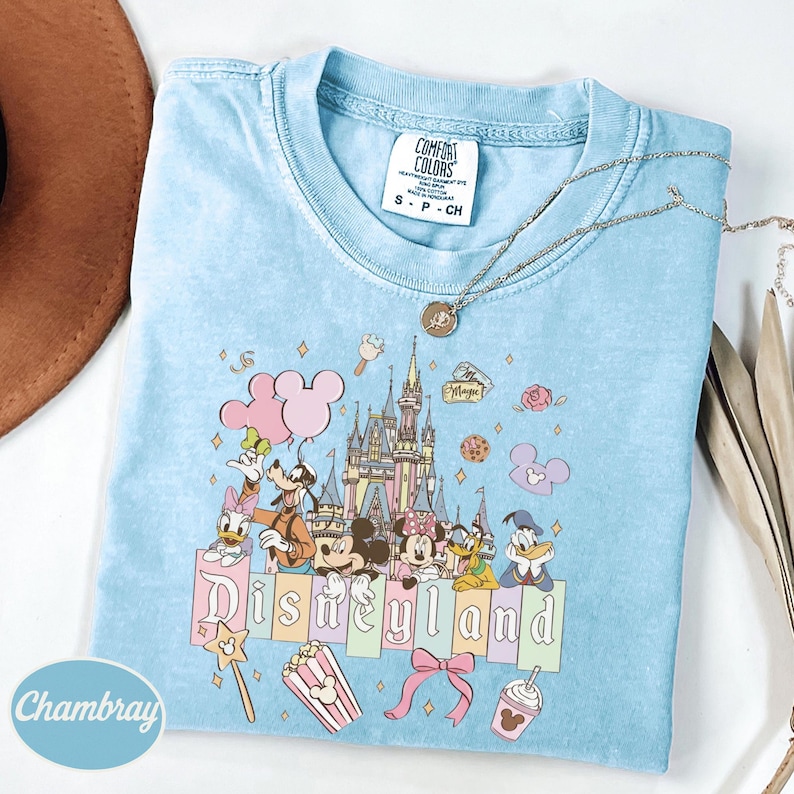 A light blue t-shirt with a Disney-themed graphic design, including Mickey Mouse, Minnie Mouse, Donald Duck, and other iconic characters, along with various Disney-related items such as popcorn and a castle.