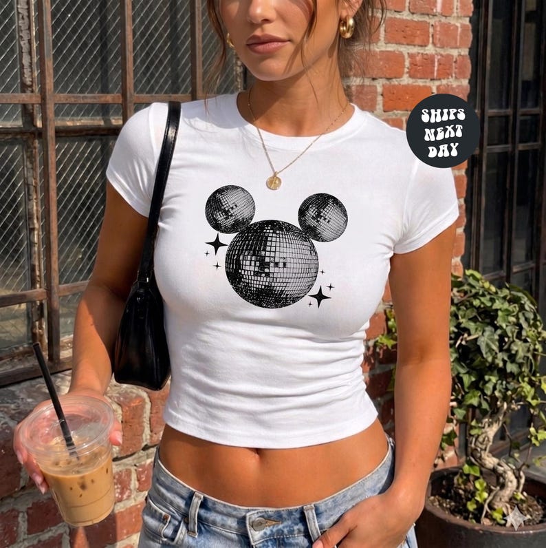 A woman wearing a white crop top with a graphic of a disco ball and stars, holding a drink and standing in front of a brick wall.