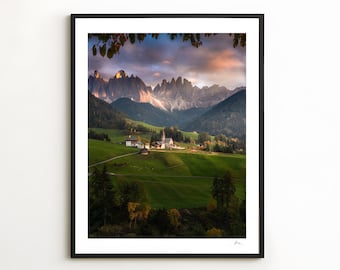 May include: A framed landscape print featuring a mountain range under a colorful sunset. The scene includes a village with a church nestled in a green valley. The print is in a black frame, ready to hang.