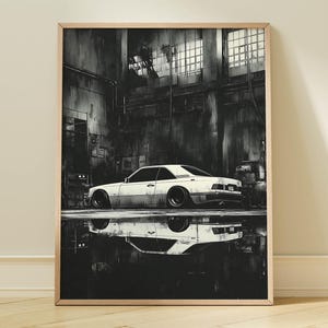 May include: A framed black and white print of a white sports car parked in a garage. The car is reflected in a puddle on the floor. The garage has a dark, industrial feel with windows and metal structures.