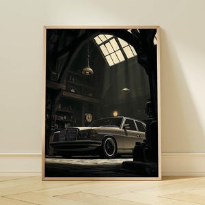 May include: A framed print depicts a classic car inside a vintage garage. The car is light-colored, with a dark grille and black tires. The garage has a high arched ceiling with a window, and the scene is lit by overhead lamps.