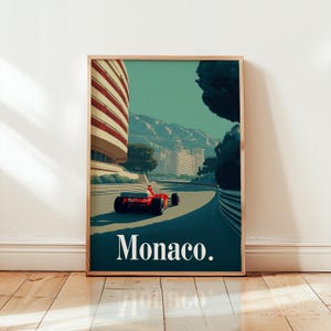 May include: Framed art print featuring a red race car speeding along a track in Monaco. The artwork showcases a building with red and white stripes, mountains, and the word "Monaco" in white text.