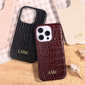 Puede incluir: Varias fundas para teléfono en tonos rosa, negro, crema y burdeos. Cada funda presenta una textura similar a la piel de cocodrilo e iniciales personalizadas. Las fundas están dispuestas sobre una superficie de madera, acompañadas de conchas marinas.