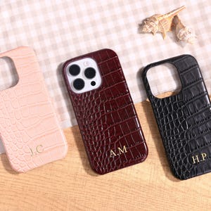 Puede incluir: Tres fundas de teléfono personalizadas en rosa, burdeos y negro, cada una con textura de piel de cocodrilo. Las fundas tienen recortes para la cámara y están grabadas con iniciales doradas: J.C., A.M. y H.P. Las fundas se muestran sobre una superficie clara.